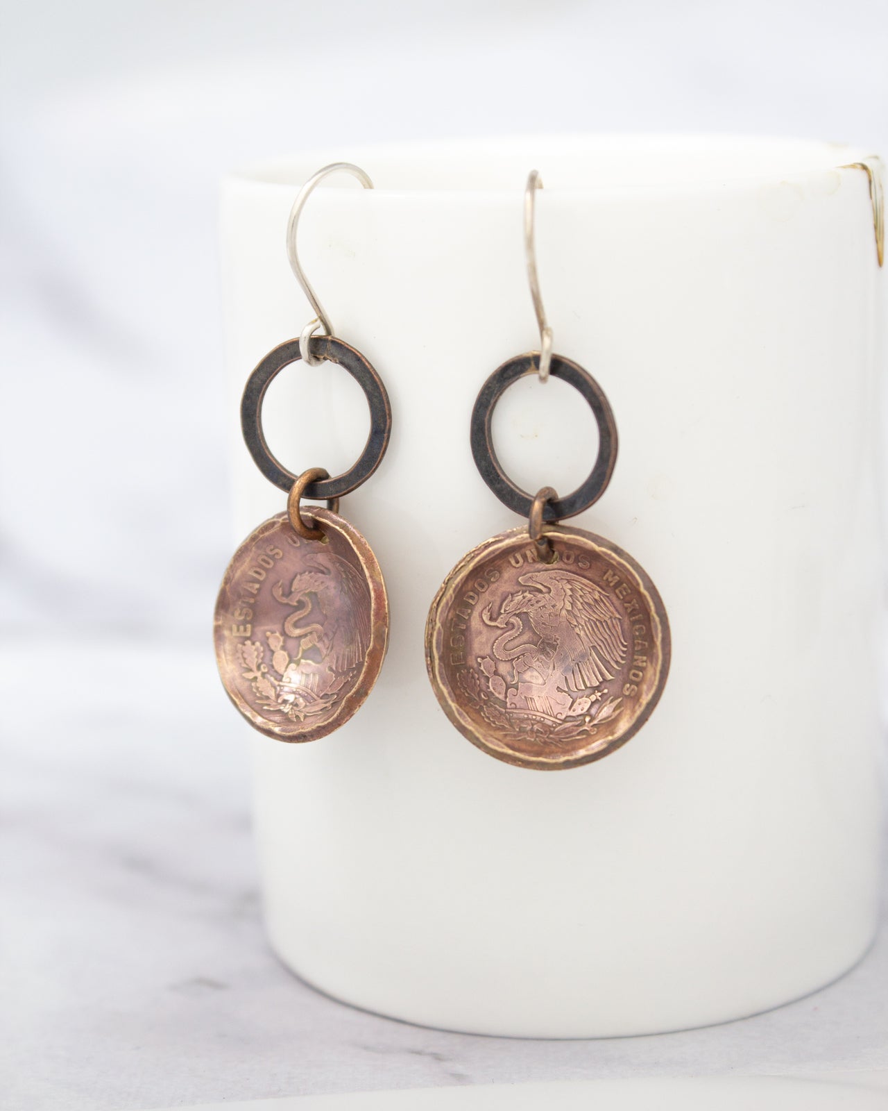 A pair of earrings made of copper-colored metal, featuring a circular design with a central motif and a smaller circular design hanging from it.