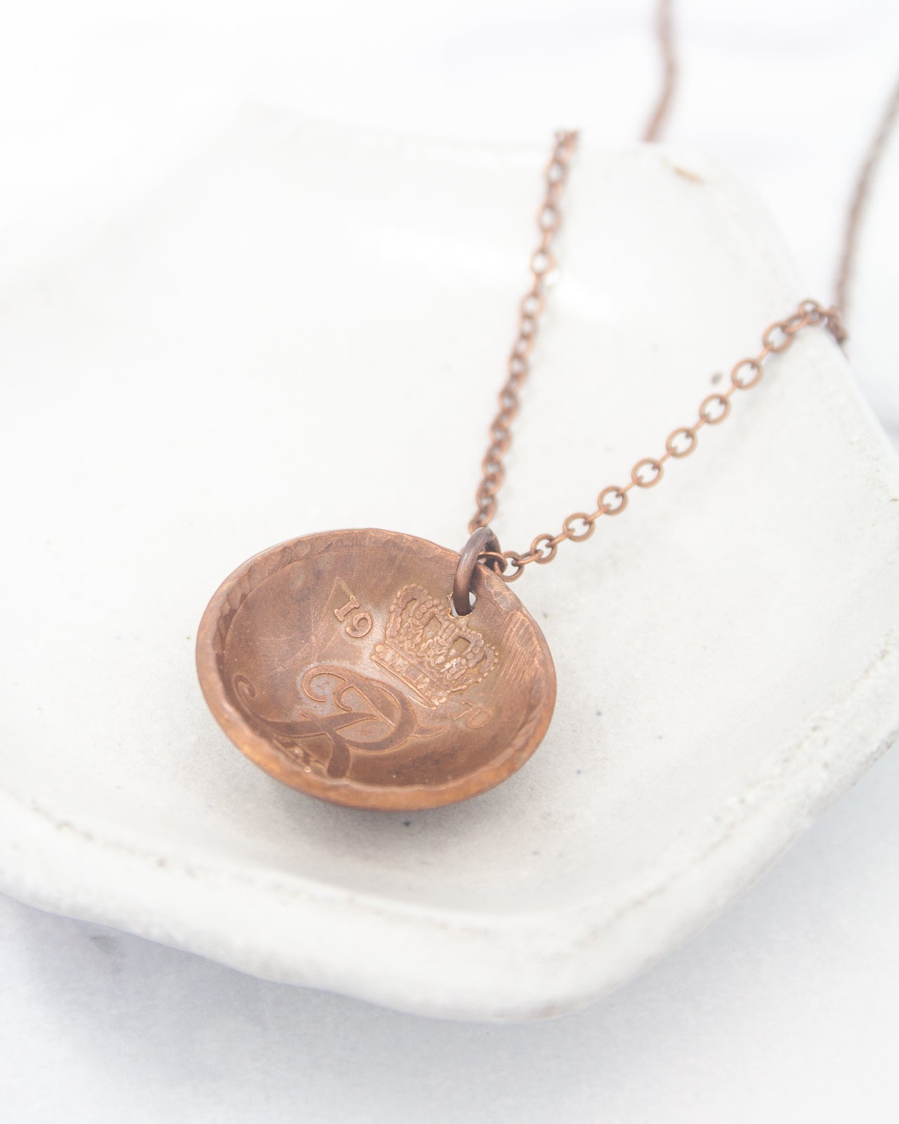 A copper-colored pendant necklace with a small engraved design on a white ceramic plate.