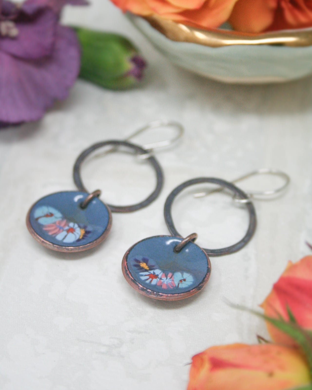 a pair of blue earrings with a floral design, placed on a white surface next to a bowl of orange flowers.