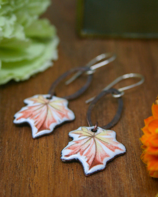 A pair of earrings with leaf-shaped designs, featuring a combination of pink and orange colors, are displayed on a wooden surface alongside some flowers.