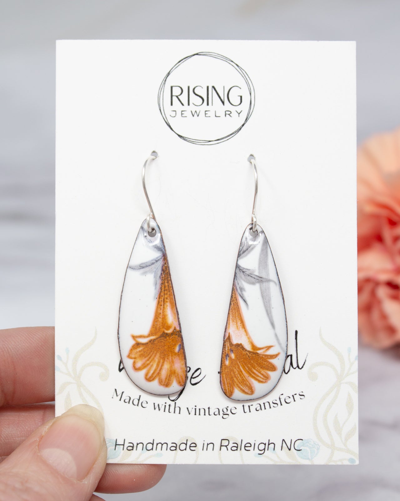 A hand holding a pair of earrings with a white and orange floral design, against a blurred background.