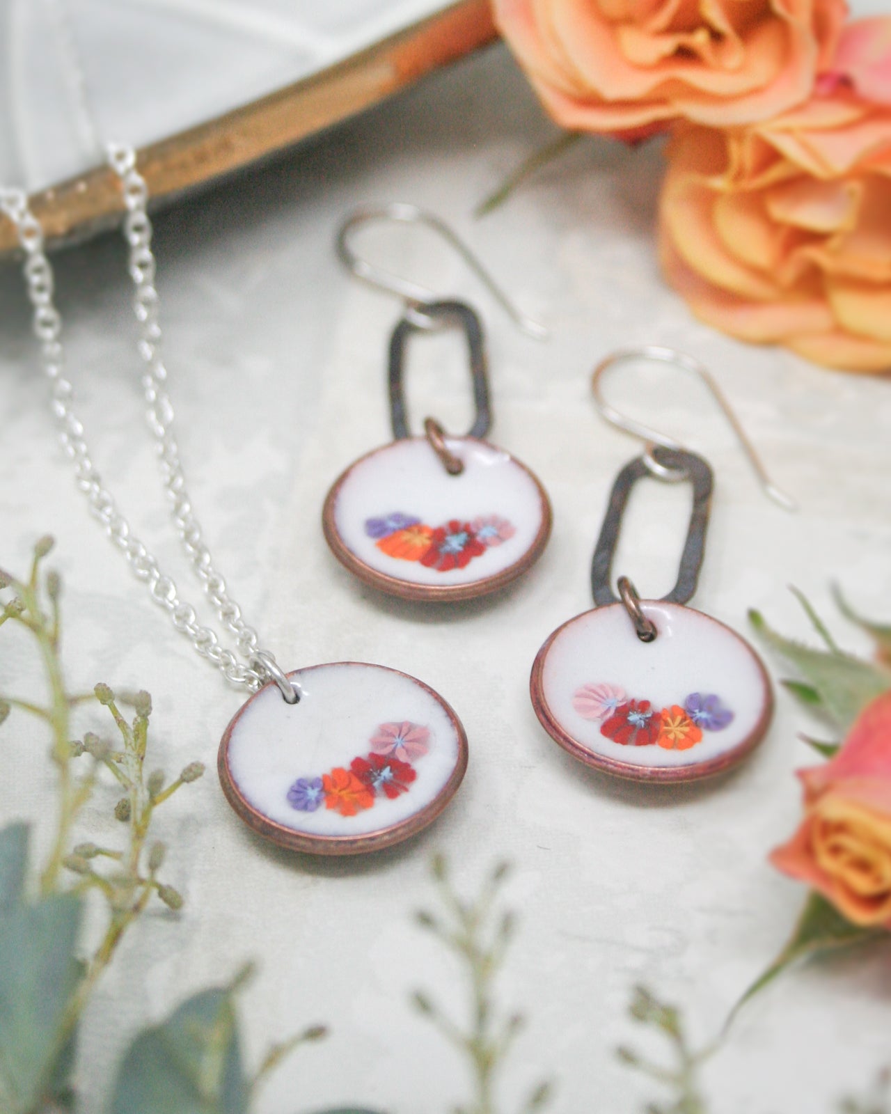 a necklace with a pendant and earrings with a floral design, placed on a surface with orange and pink flowers.