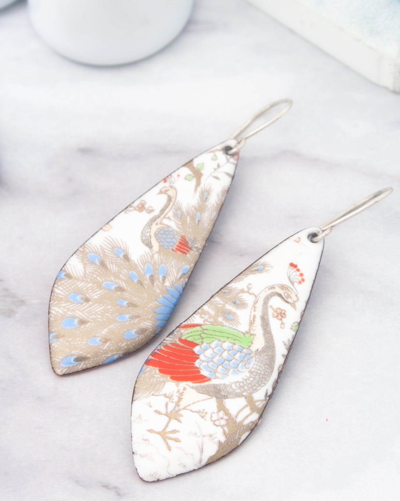 two ornate earrings with intricate designs, including a peacock and a bird, resting on a marble surface.