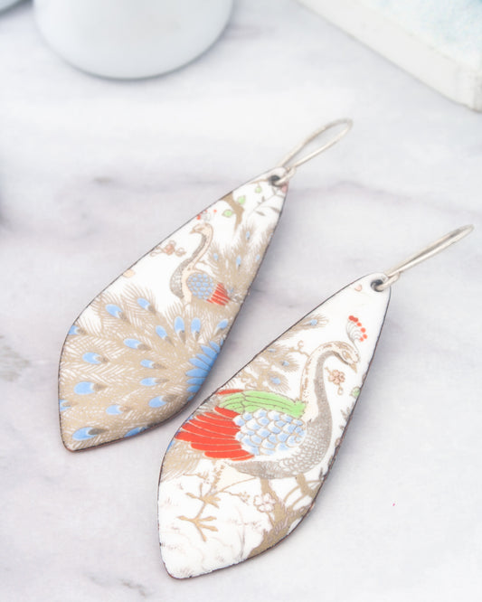 two ornate earrings with intricate designs, including a peacock and a bird, resting on a marble surface.