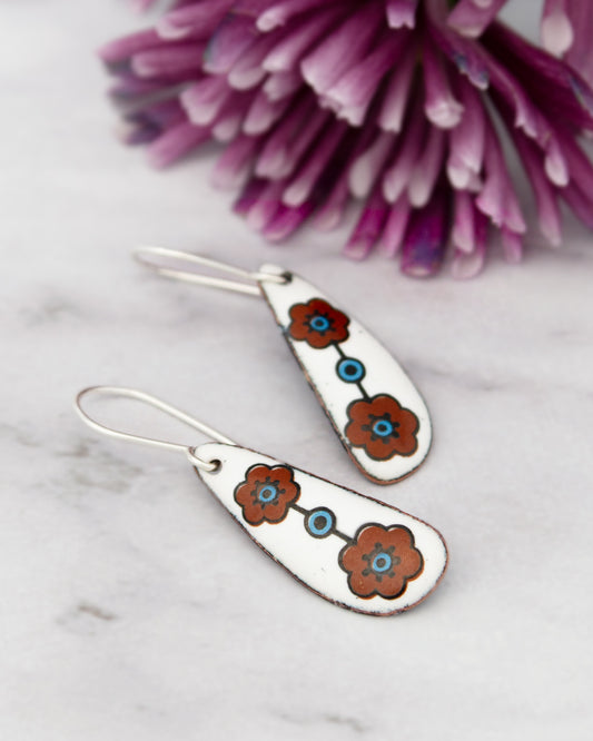 A pair of white earrings with blue flower designs on a marble surface, with a purple flower in the background.