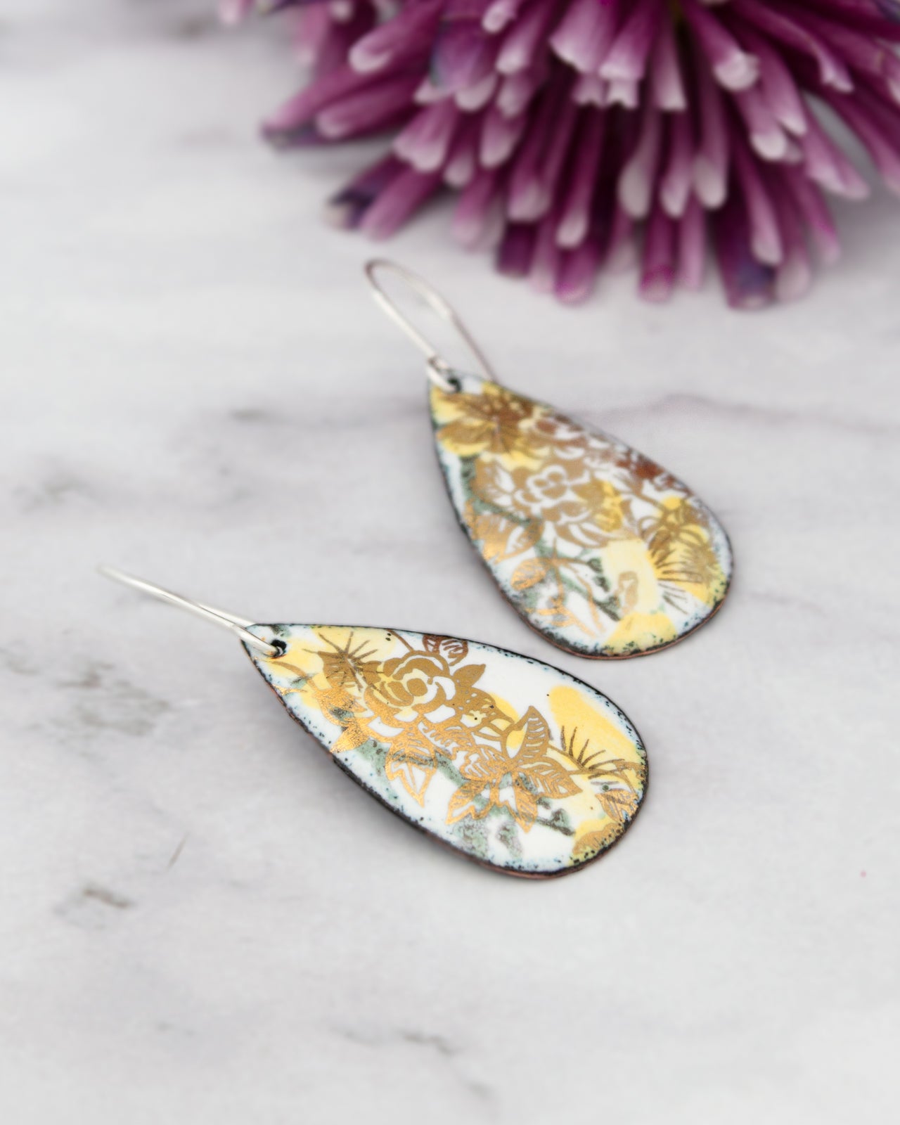 a pair of earrings with a floral design, placed on a marble surface next to a purple flower.