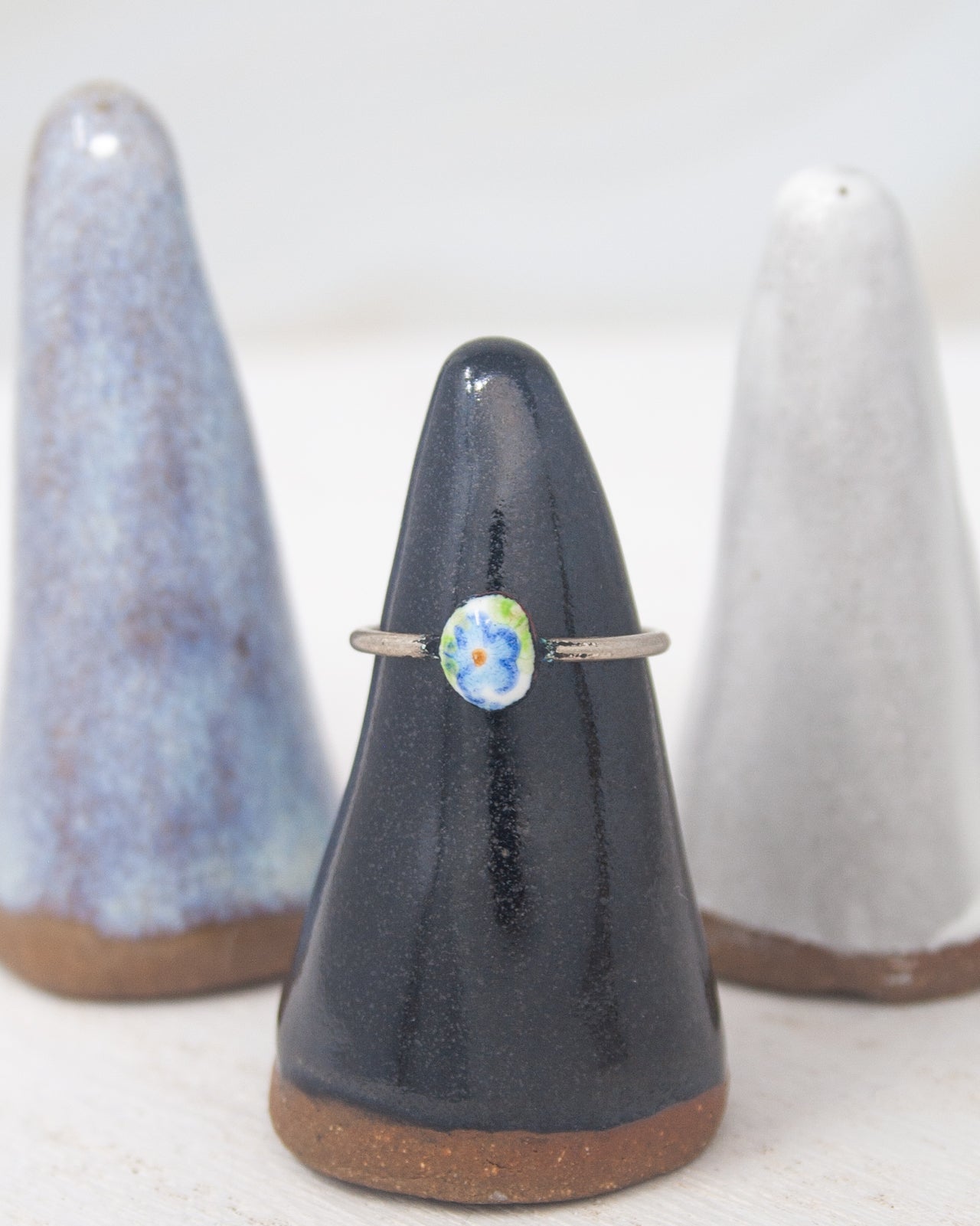 A ring with a blue and green flower design is displayed on a black stand, with two white cone-shaped objects in the background.