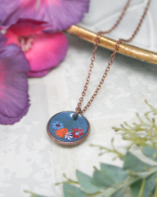 A necklace with a blue pendant featuring colorful flowers is displayed on a white surface, accompanied by a purple flower and some greenery.