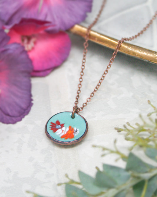 A necklace with a circular pendant featuring a floral design is displayed on a white surface, surrounded by purple and green flowers.