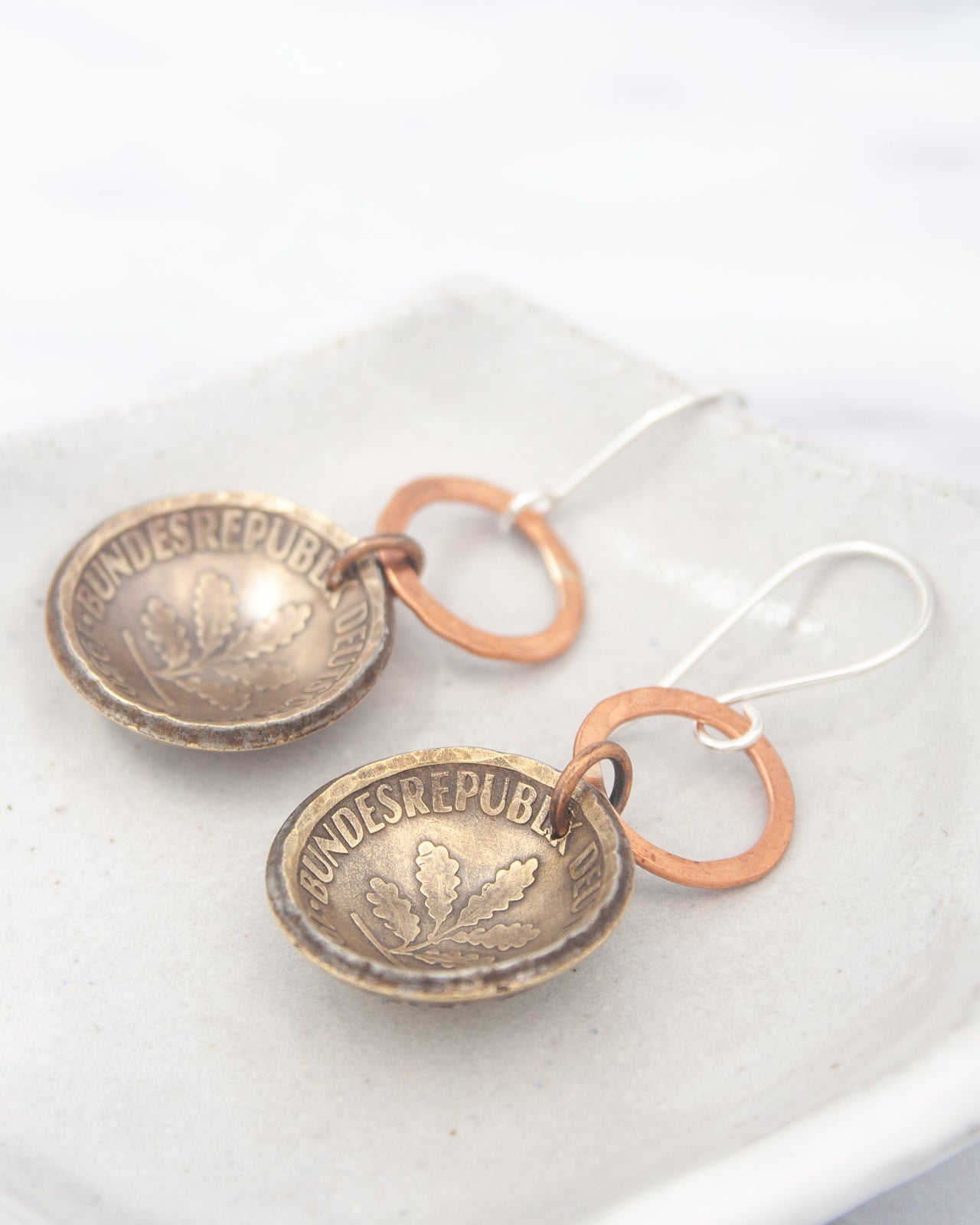 a pair of earrings made of metal, featuring a design that resembles a coin. The earrings are placed on a white surface, and they appear to be made of copper and brass materials.