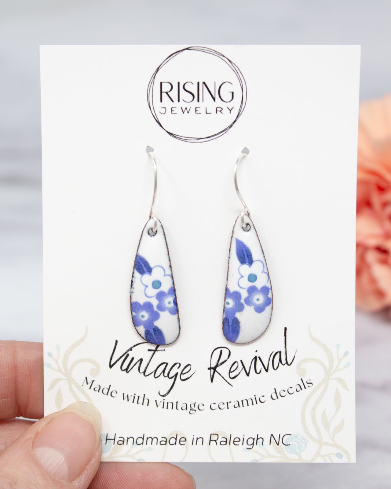 A hand holding a pair of earrings with a card that displays the earrings and the company's name.
