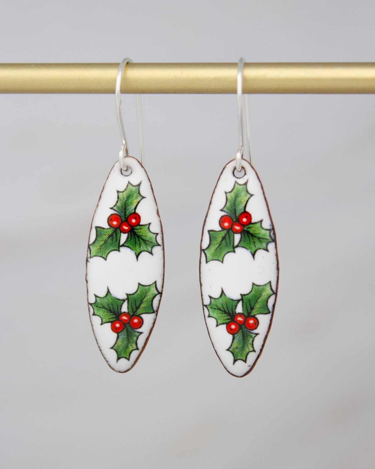 A pair of white earrings with green holly leaves and red berries on a gold bar.