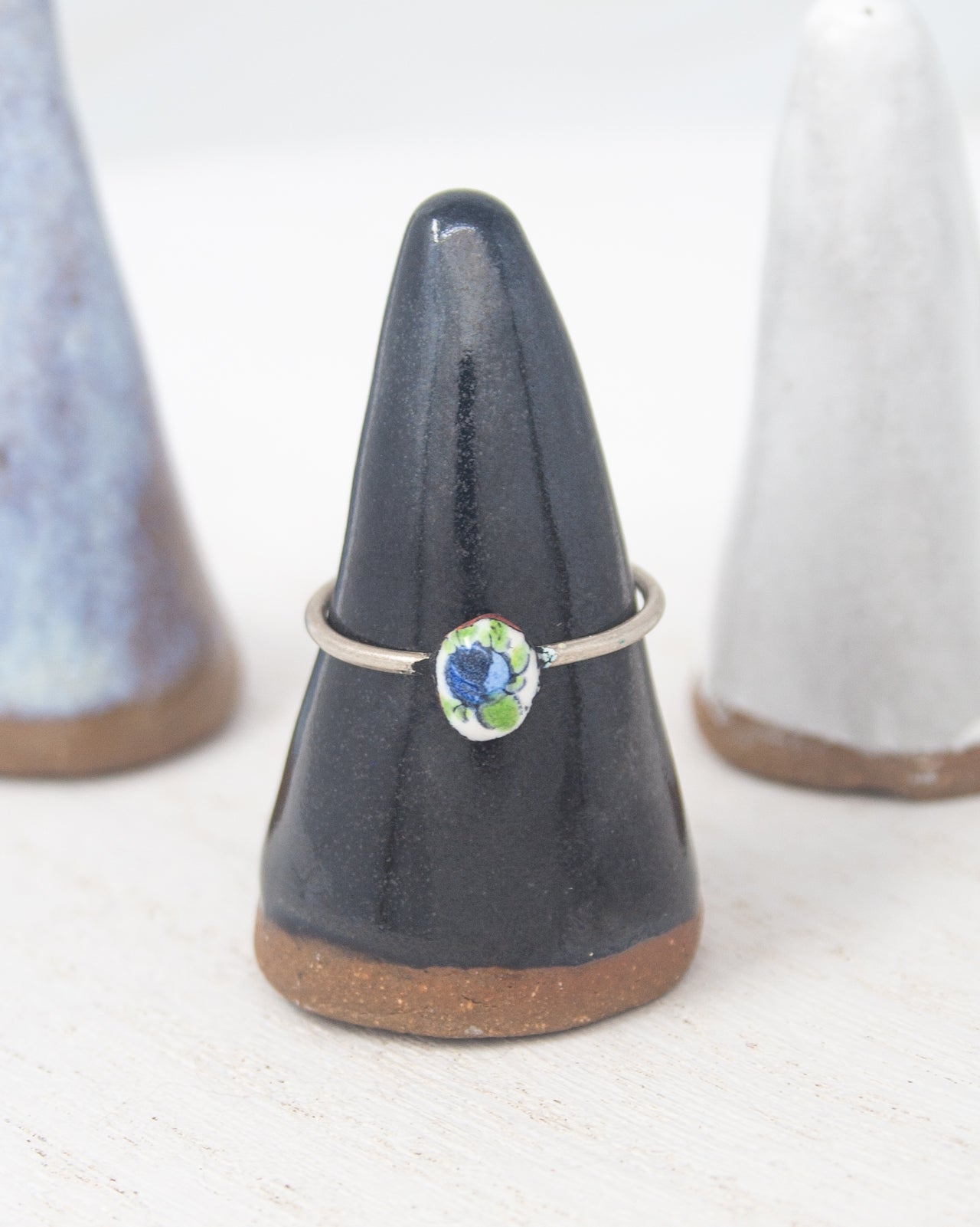 A silver ring with a blue and green floral design is displayed on a black ceramic stand, with two white ceramic stands in the background.