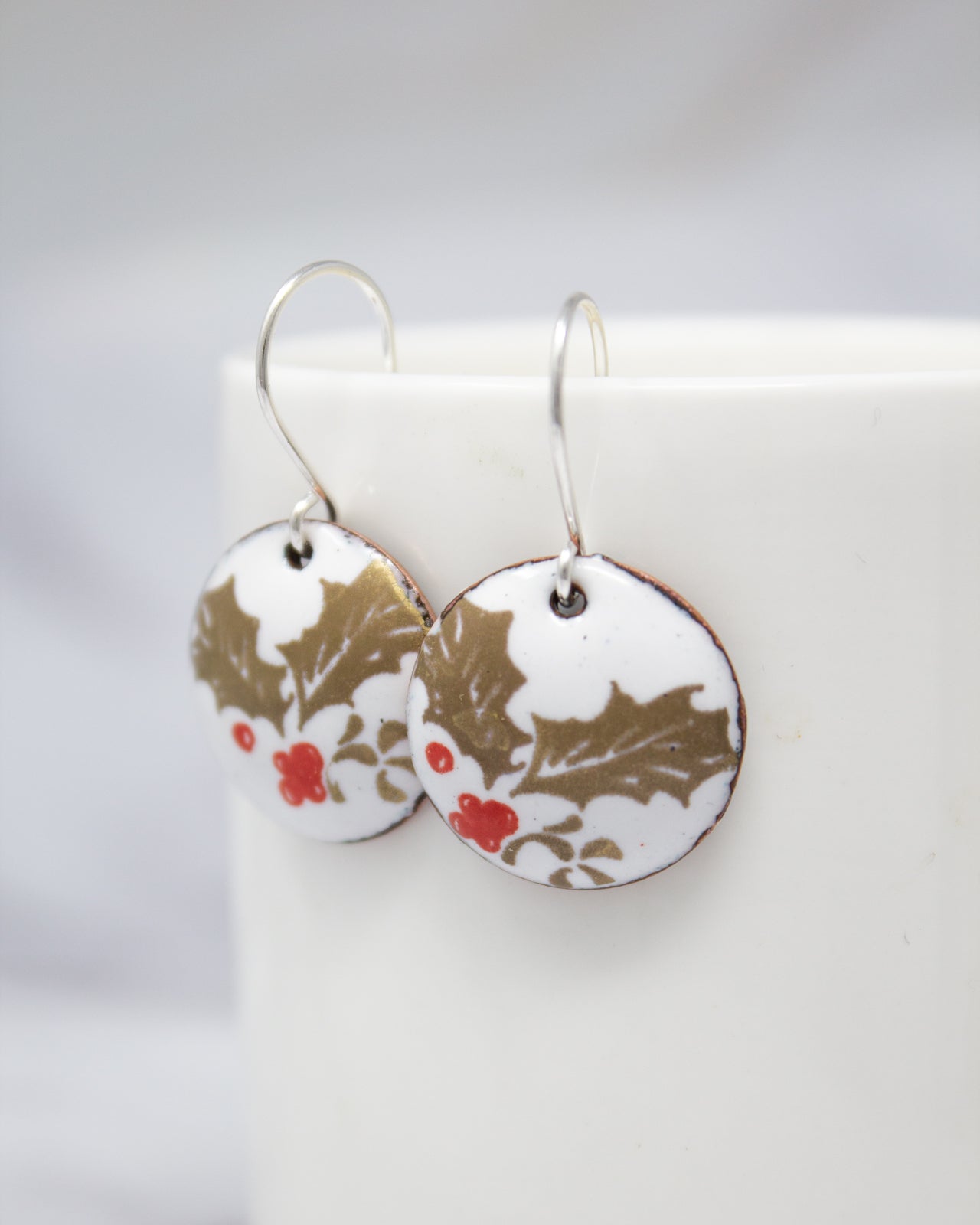 A pair of round earrings with a floral design, featuring red berries and holly leaves, are displayed on a white surface.