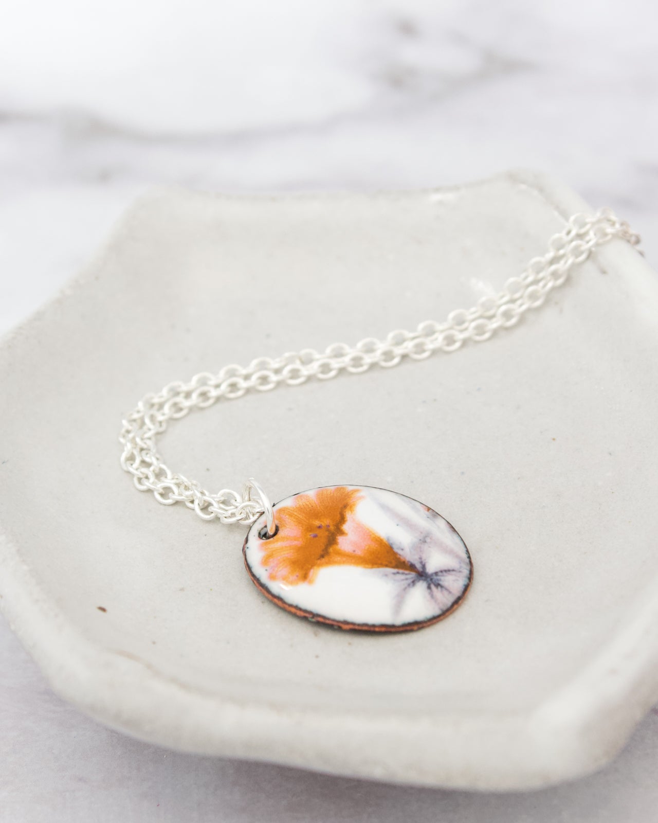 A necklace with a circular pendant featuring a floral design in orange and white colors, resting on a white plate.