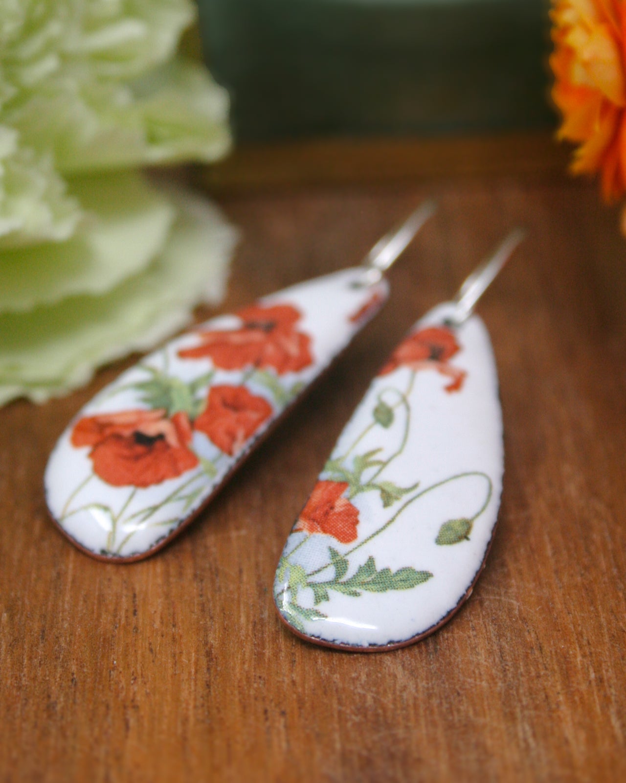 A pair of white earrings with red flowers and green leaves, placed on a wooden surface.