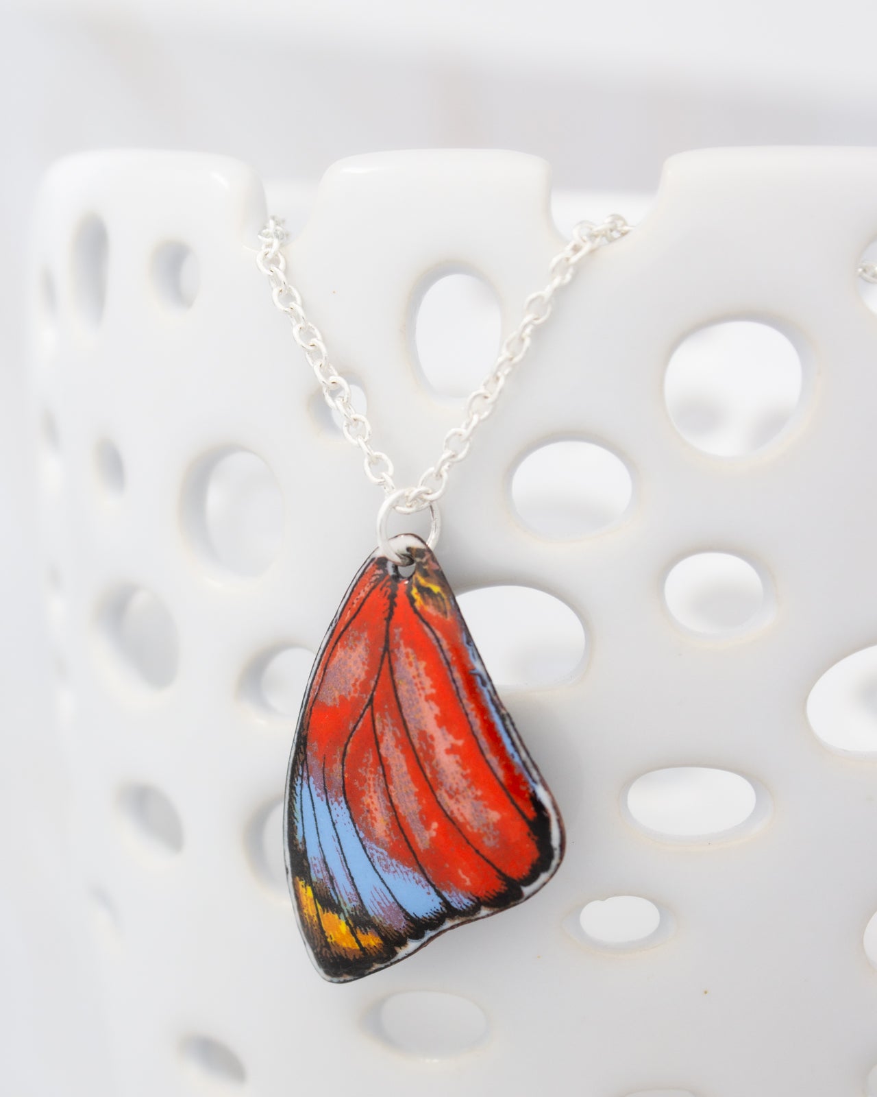 A colorful butterfly wing pendant is hanging from a chain against a white background.