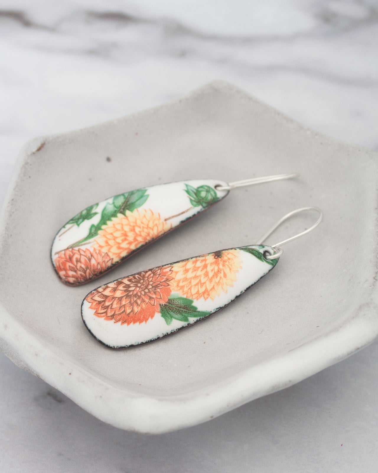 A pair of earrings with a floral design, featuring orange and green flowers, are displayed on a white plate.