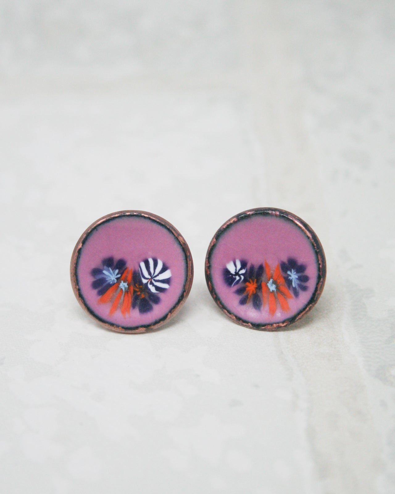 two round earrings with a floral design, featuring purple and orange flowers, resting on a white surface.
