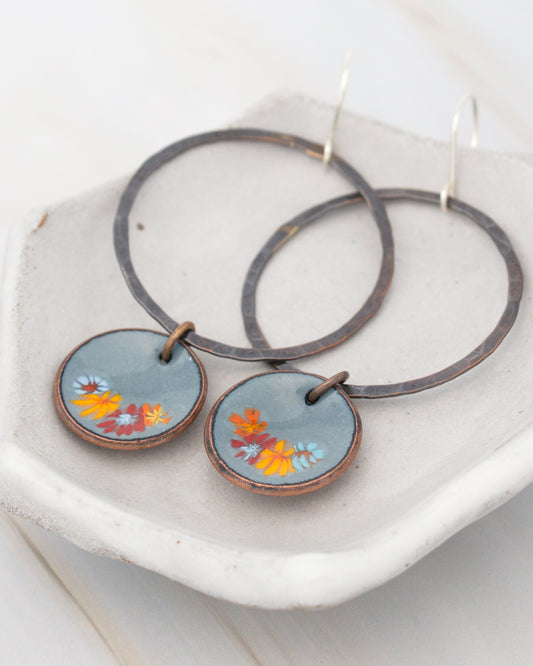 A pair of round earrings with blue and orange floral designs, hanging from a pair of black metal rings.