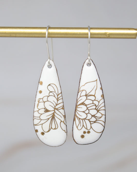 A pair of white earrings with gold floral designs hanging from a gold bar.