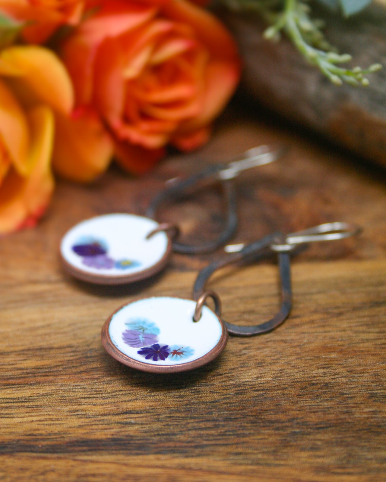 A pair of round earrings with floral designs on a wooden surface, with a blurred background featuring orange and red roses.