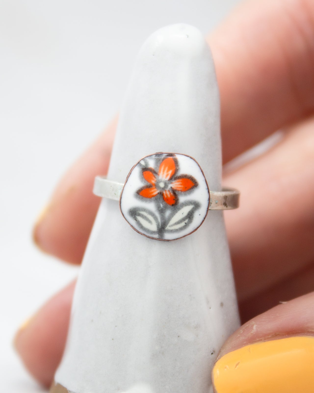 A close-up image of a person's hand wearing a ring with a flower design on it, placed on a white stand.
