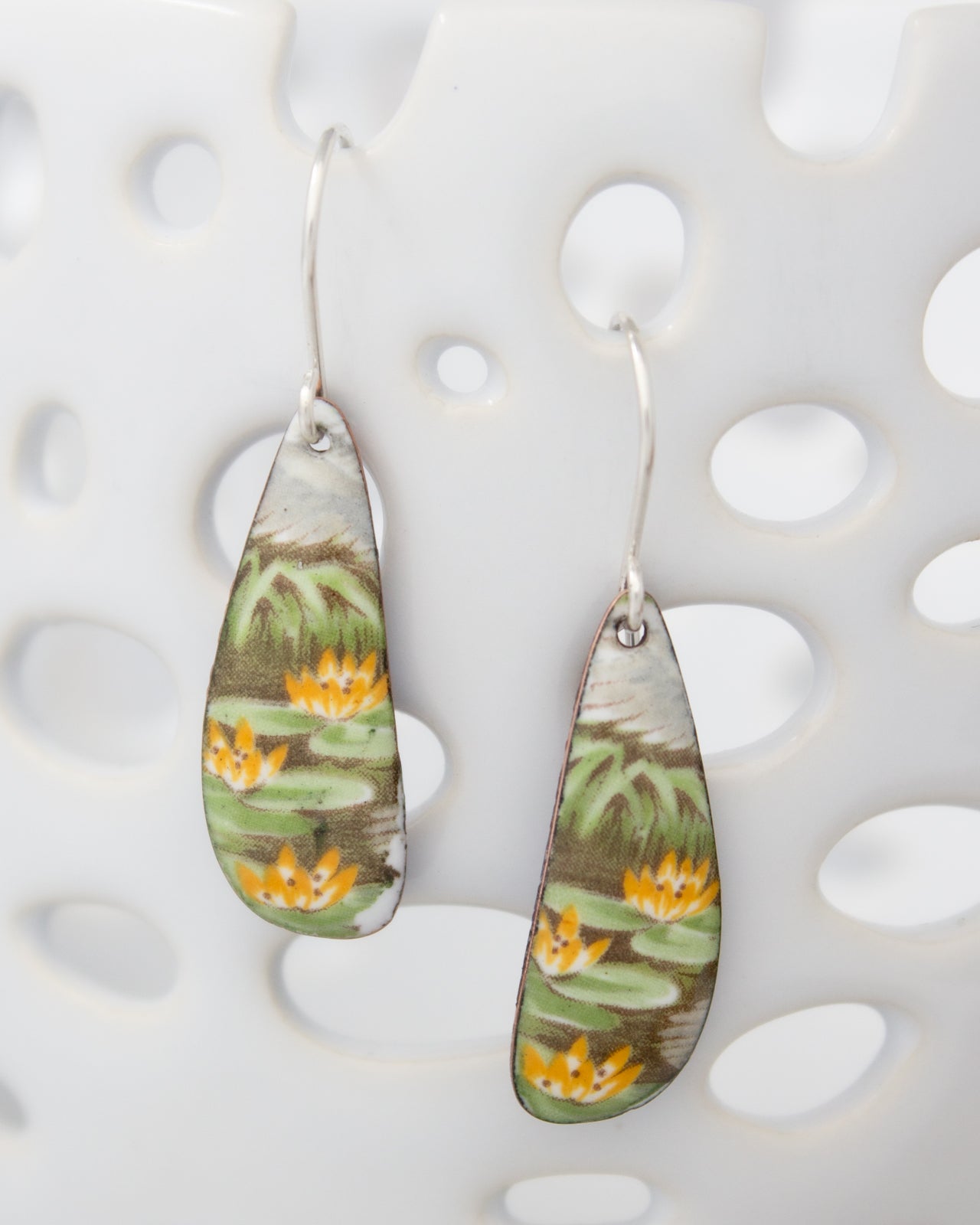 A pair of earrings with a floral design, featuring green leaves and yellow flowers, hanging from a hook against a white background.