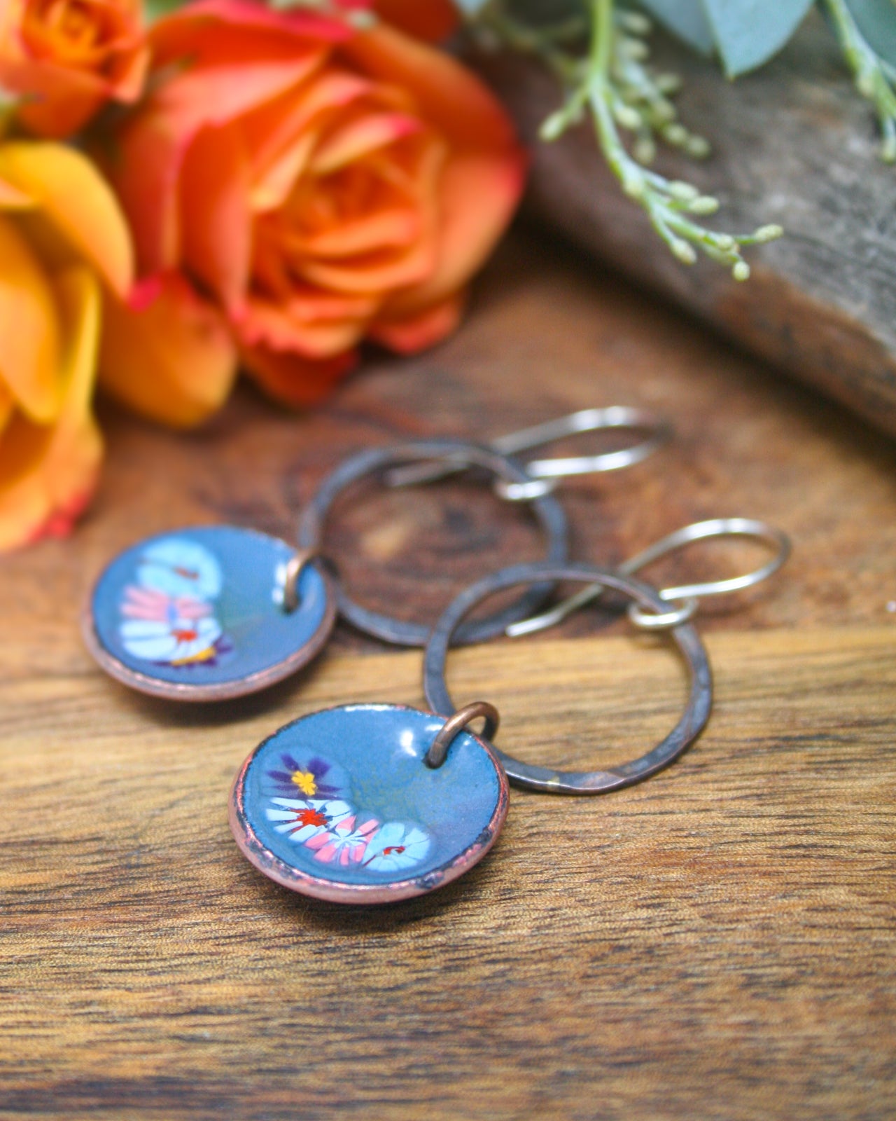 A pair of blue earrings with floral designs rests on a wooden surface, accompanied by a bouquet of orange and yellow roses.