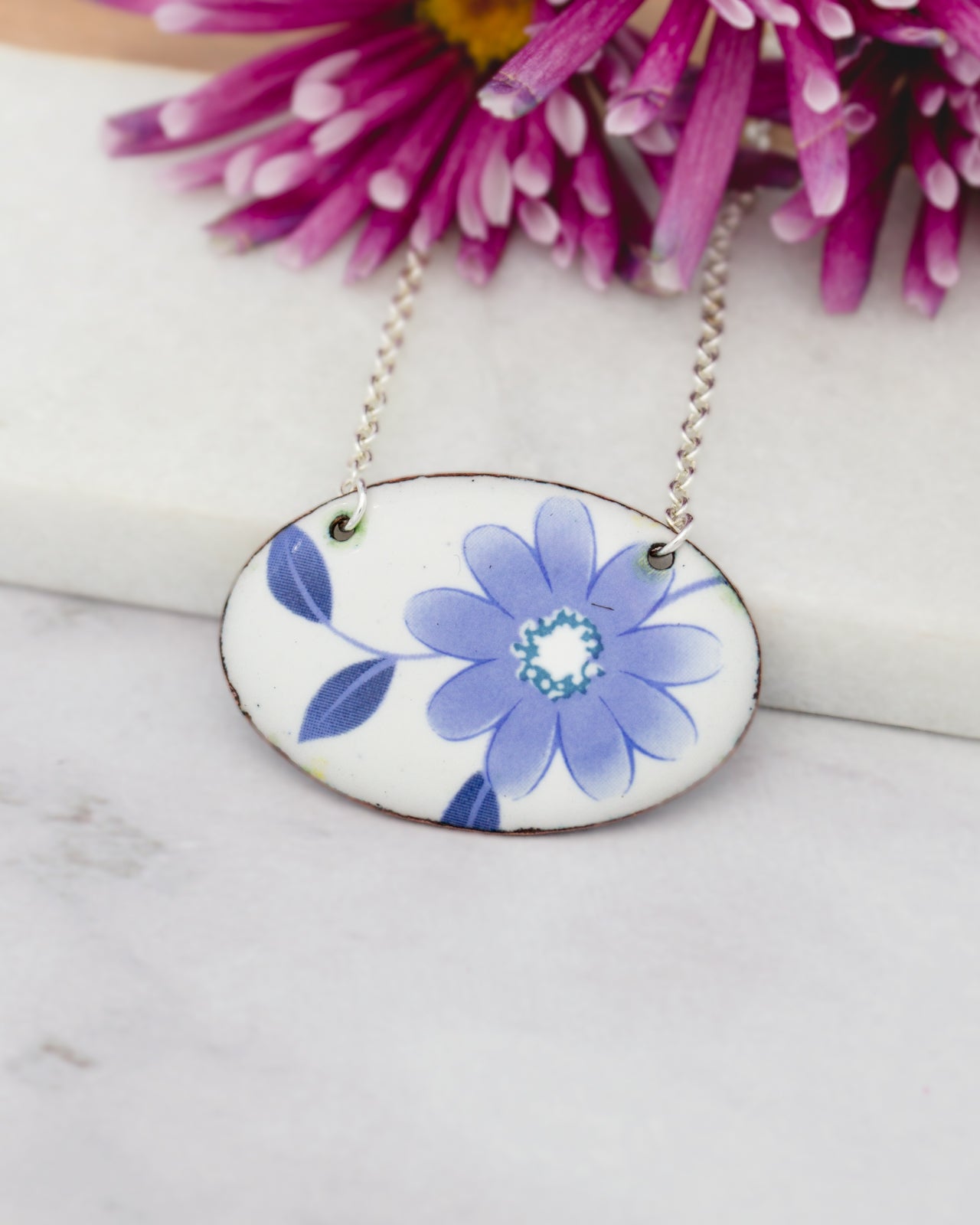 A necklace with a blue and white floral design on an oval-shaped pendant, resting on a white surface with a purple flower in the background.
