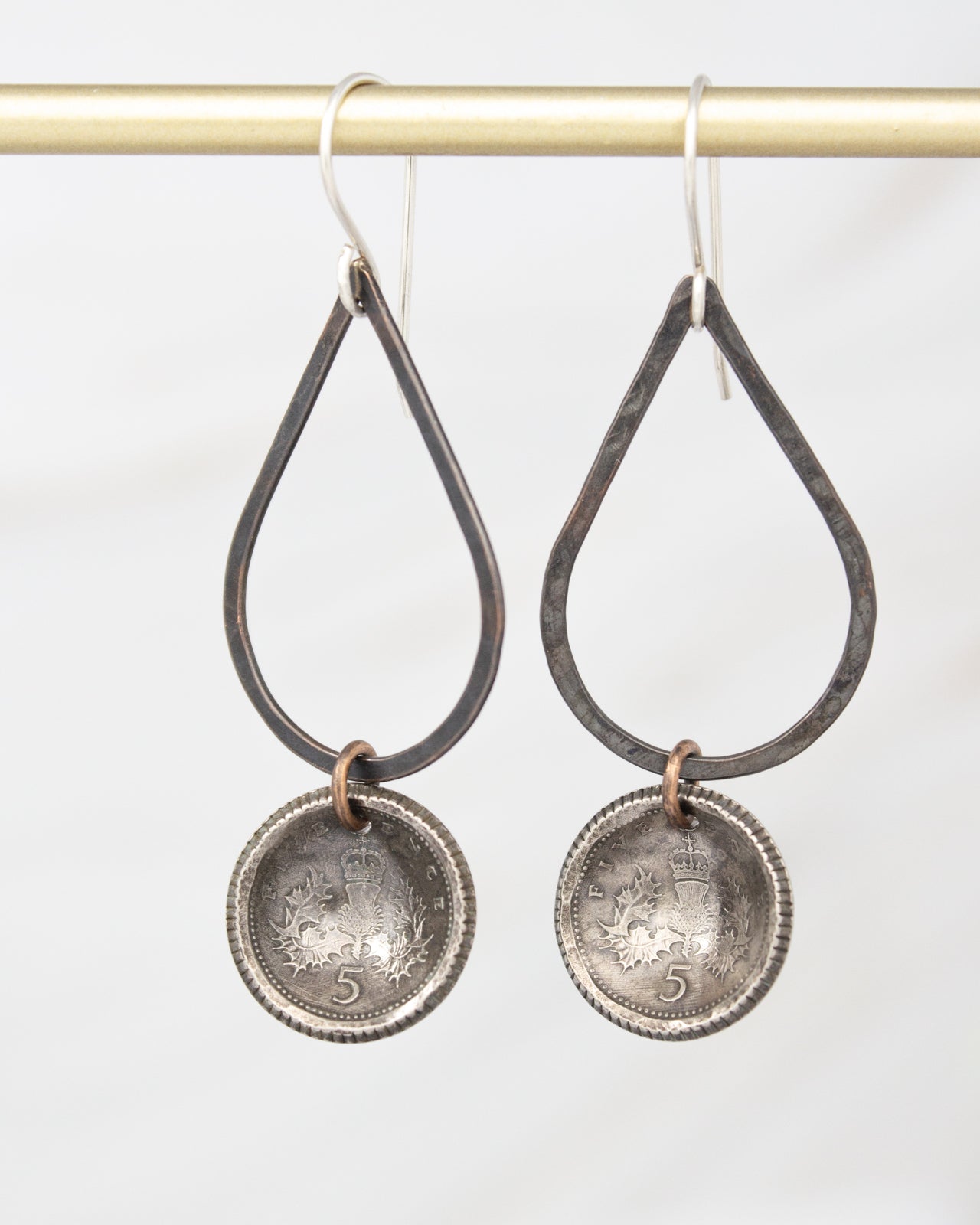 A pair of earrings hanging from a gold-colored metal hook. The earrings have a circular metal pendant with an embossed design, and they are suspended from a gold-colored metal hook.