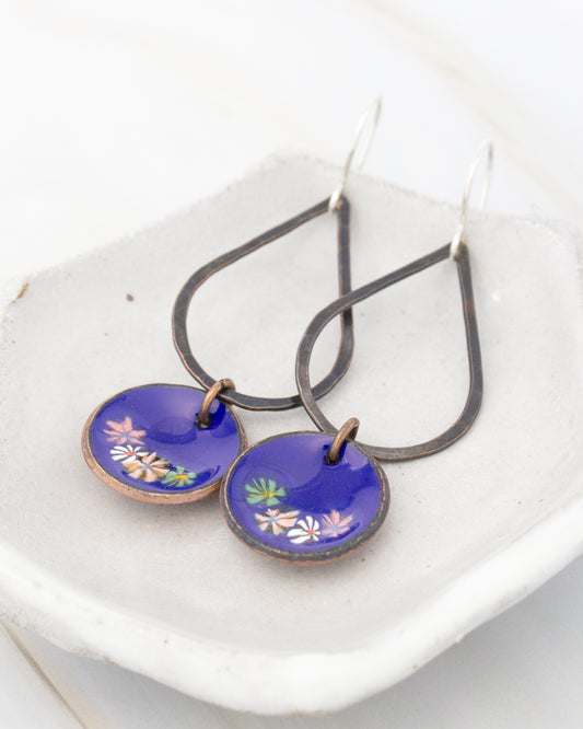 A pair of earrings with a floral design on the blue pendants, resting on a white surface.