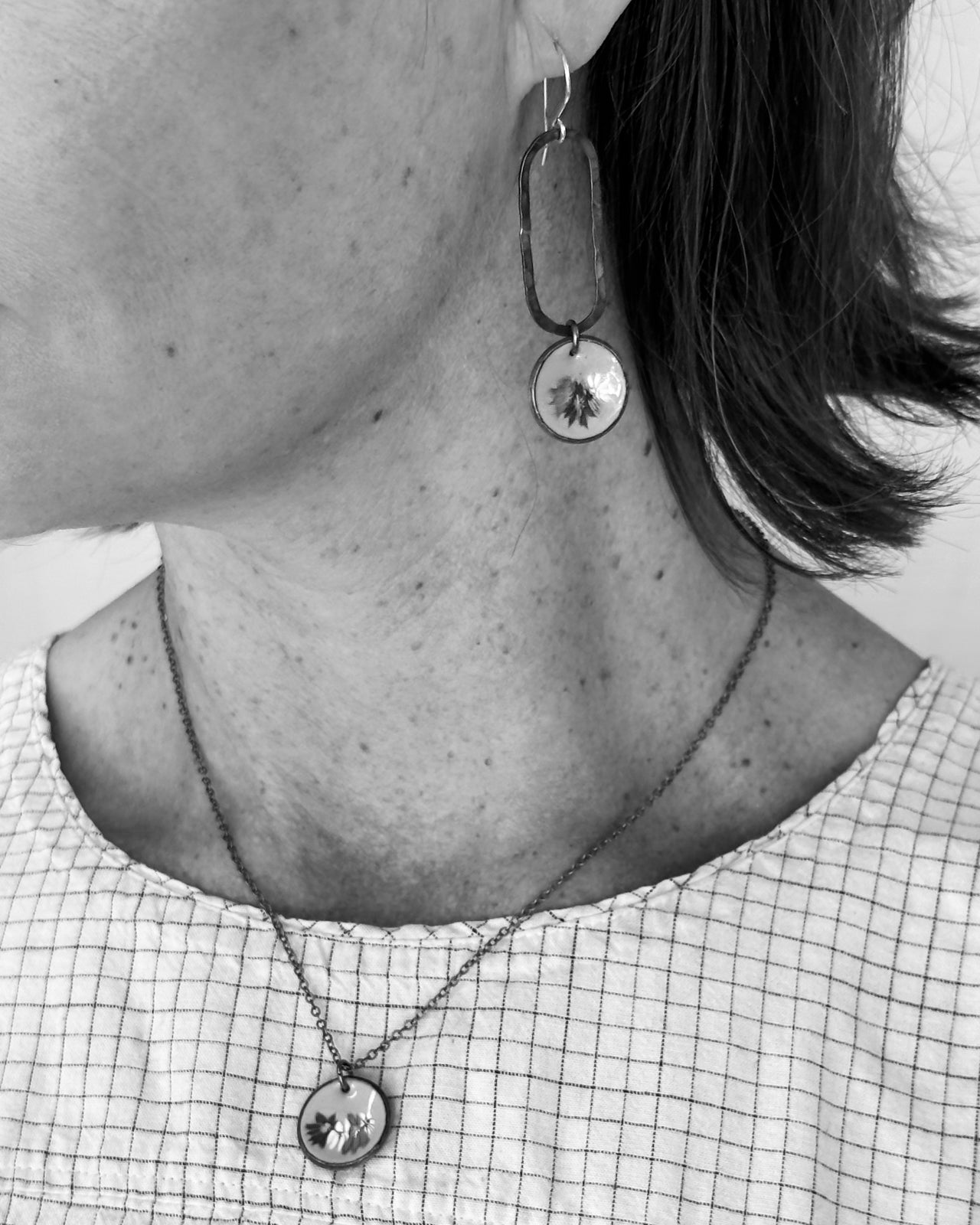 A close-up black and white image of a person's neck and earrings, with a necklace and a pendant visible.