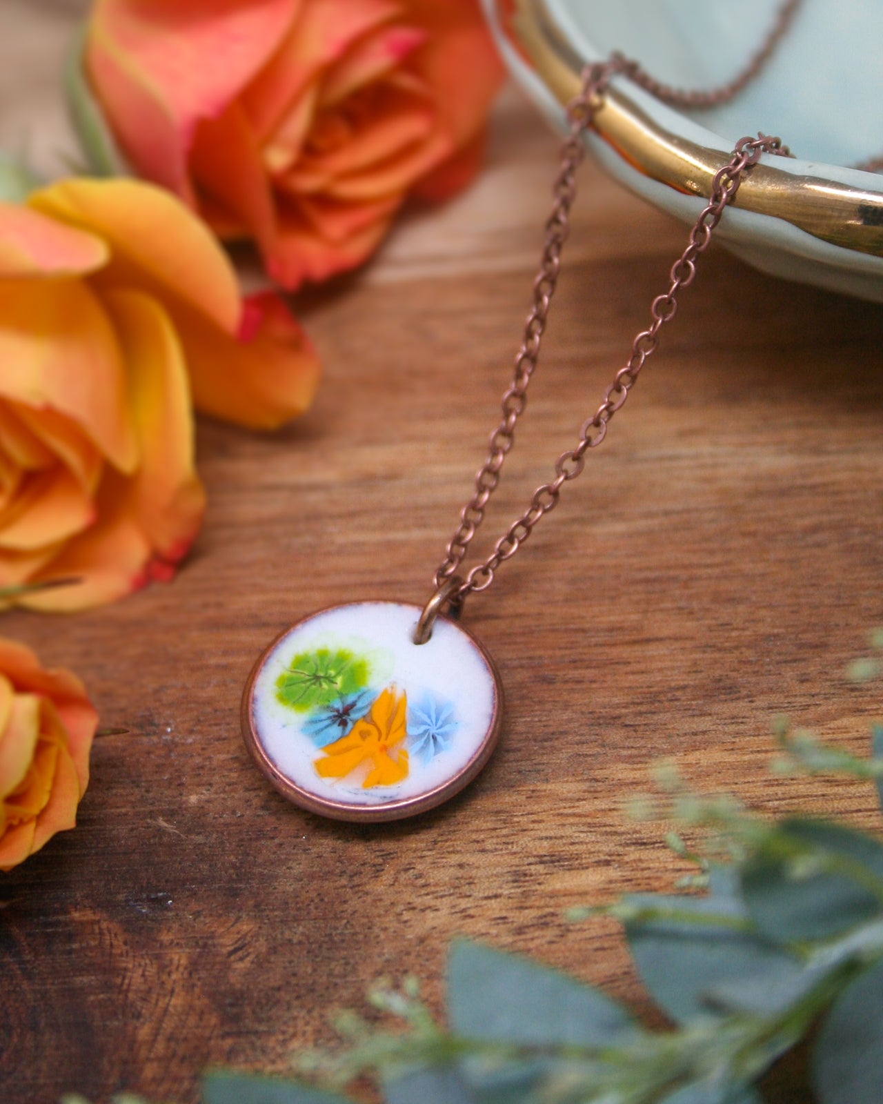 a necklace with a pendant, surrounded by a variety of flowers, including roses and greenery, on a wooden surface.
