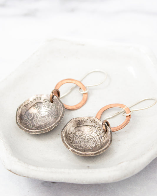A pair of silver earrings with a circular design and the word "TWENTY" engraved on them, displayed on a white plate.