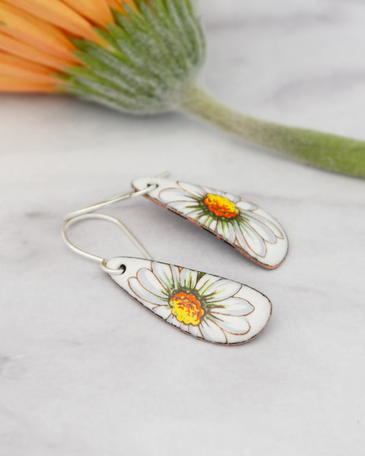 A pair of earrings with white and orange flower designs, placed on a marble surface next to a green flower stem.