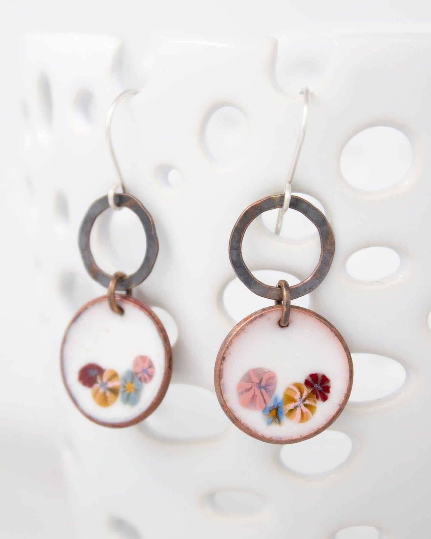 A pair of earrings with a floral design on the circular pendants, hanging from a hook against a white background.