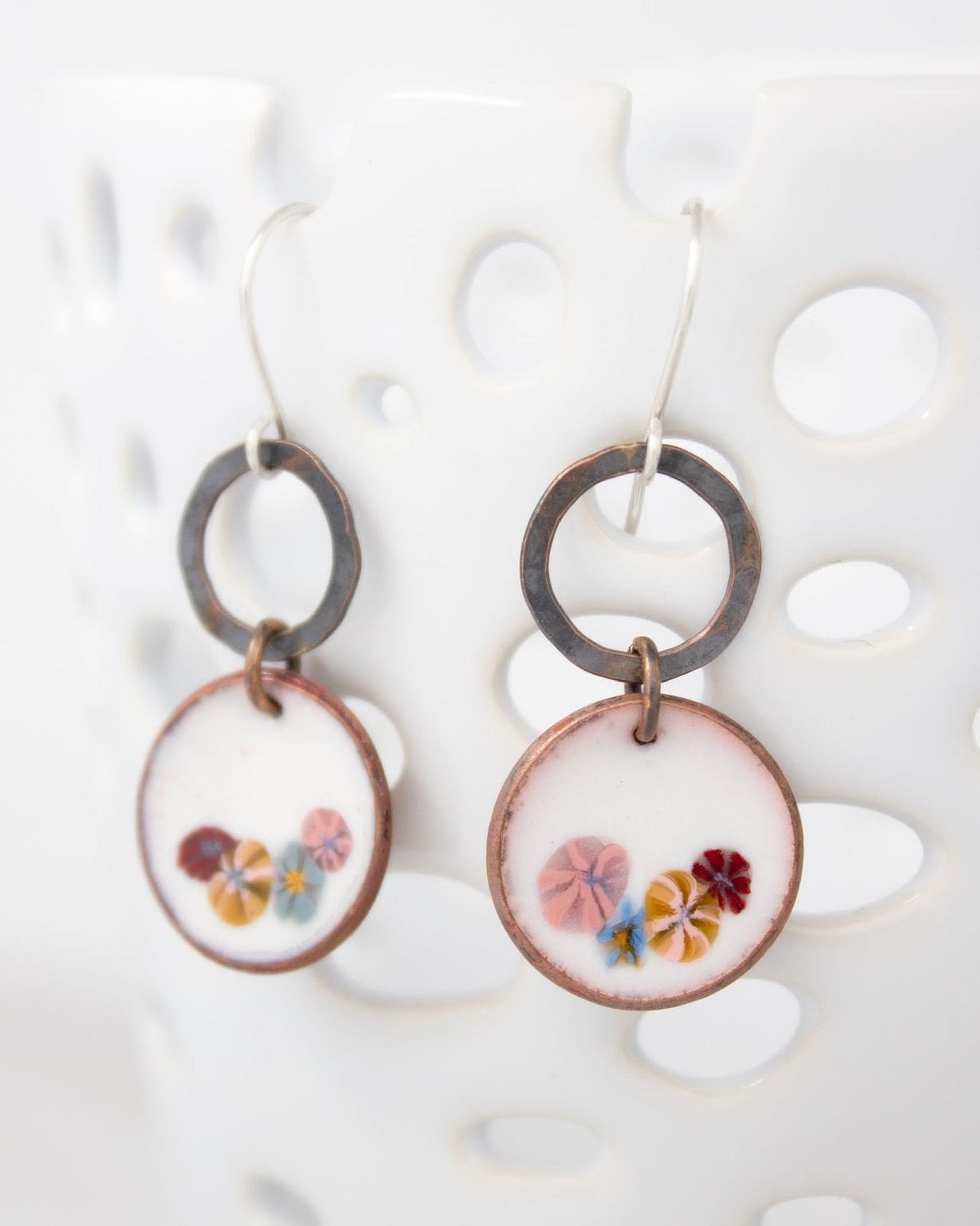 A pair of earrings with a floral design on the circular pendants, hanging from a hook against a white background.
