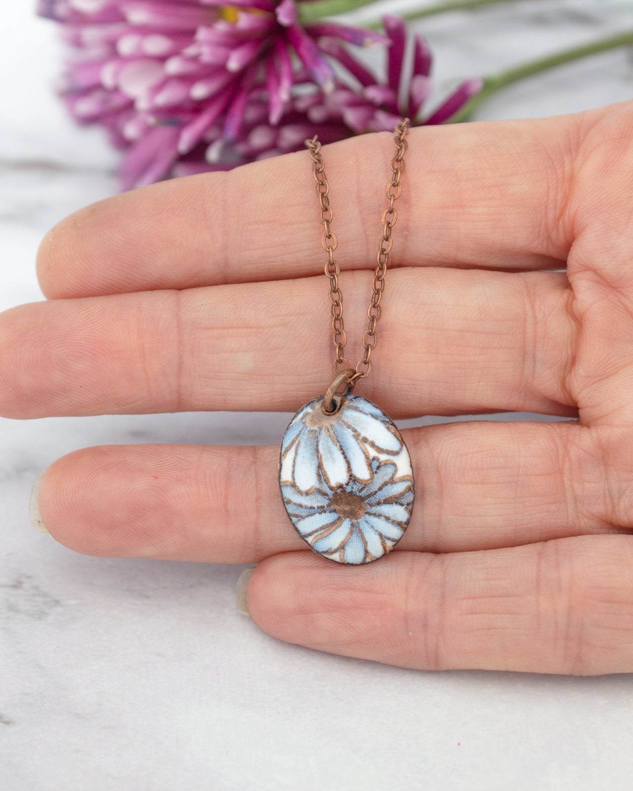 A hand holding a delicate necklace with a pendant featuring a blue flower design.