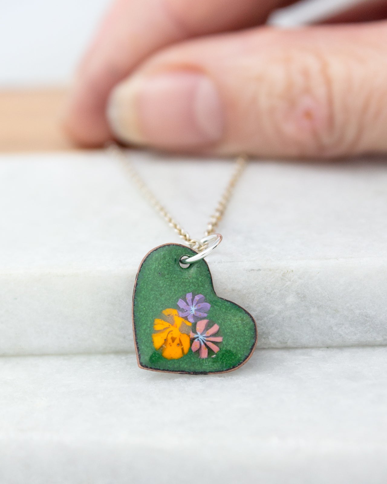 A hand is holding a green heart-shaped pendant with colorful flowers on it, resting on a white surface.