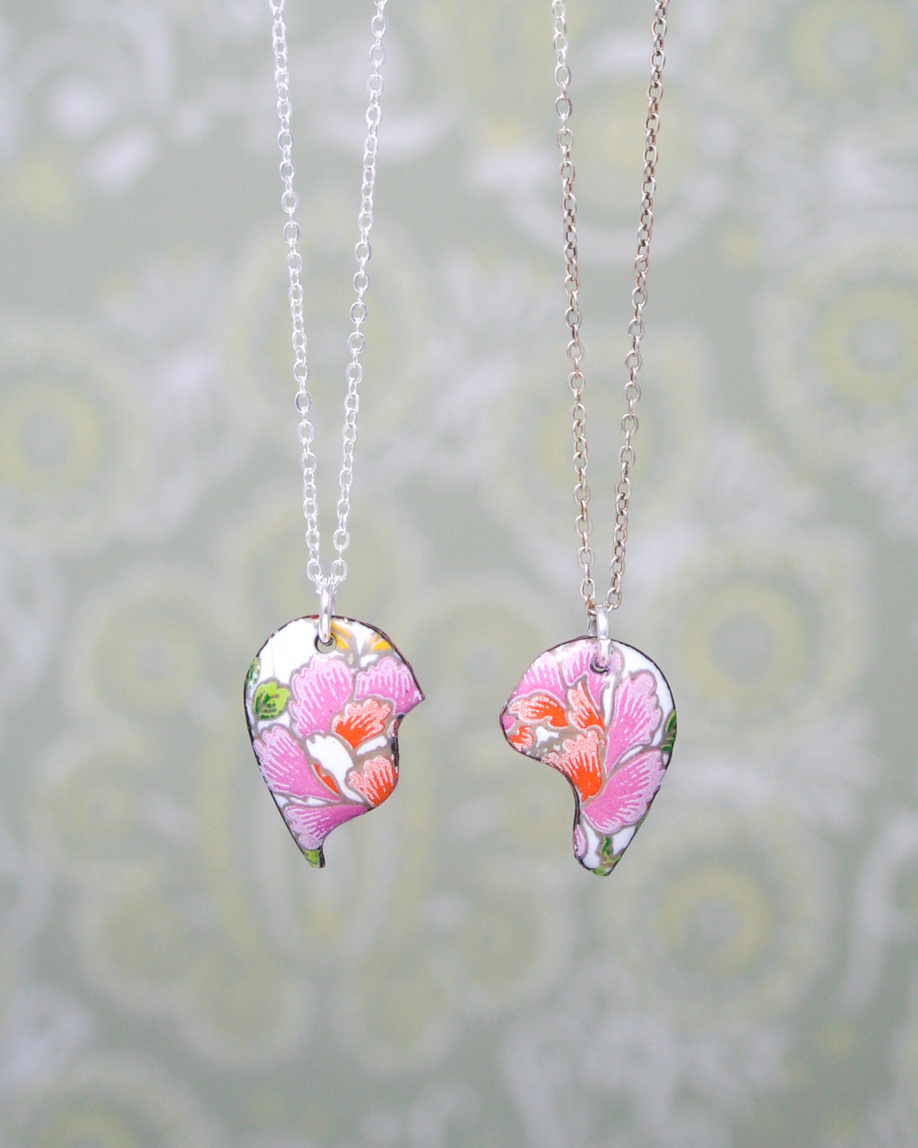two pendant necklaces with floral designs, hanging against a patterned background.