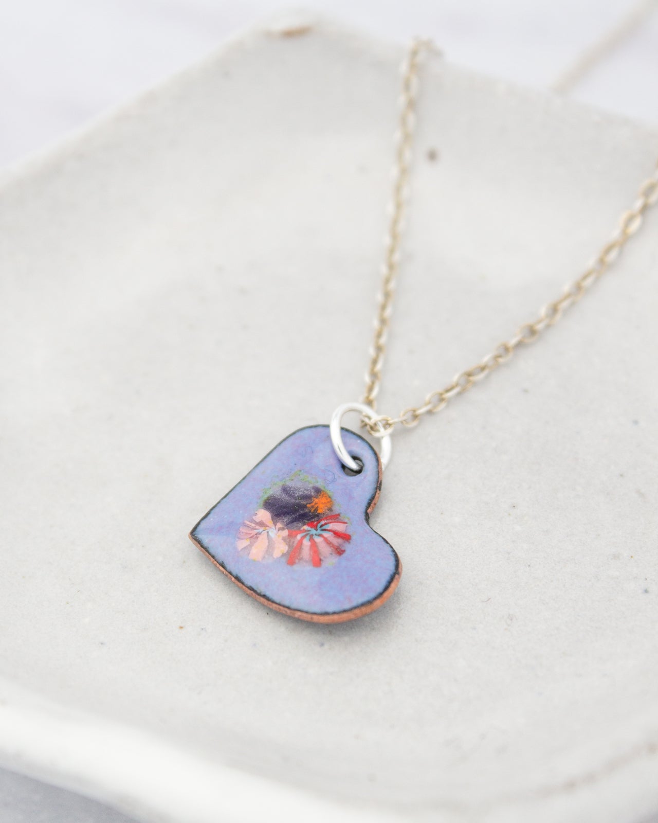 A heart-shaped pendant with a floral design is displayed on a white surface, attached to a chain.