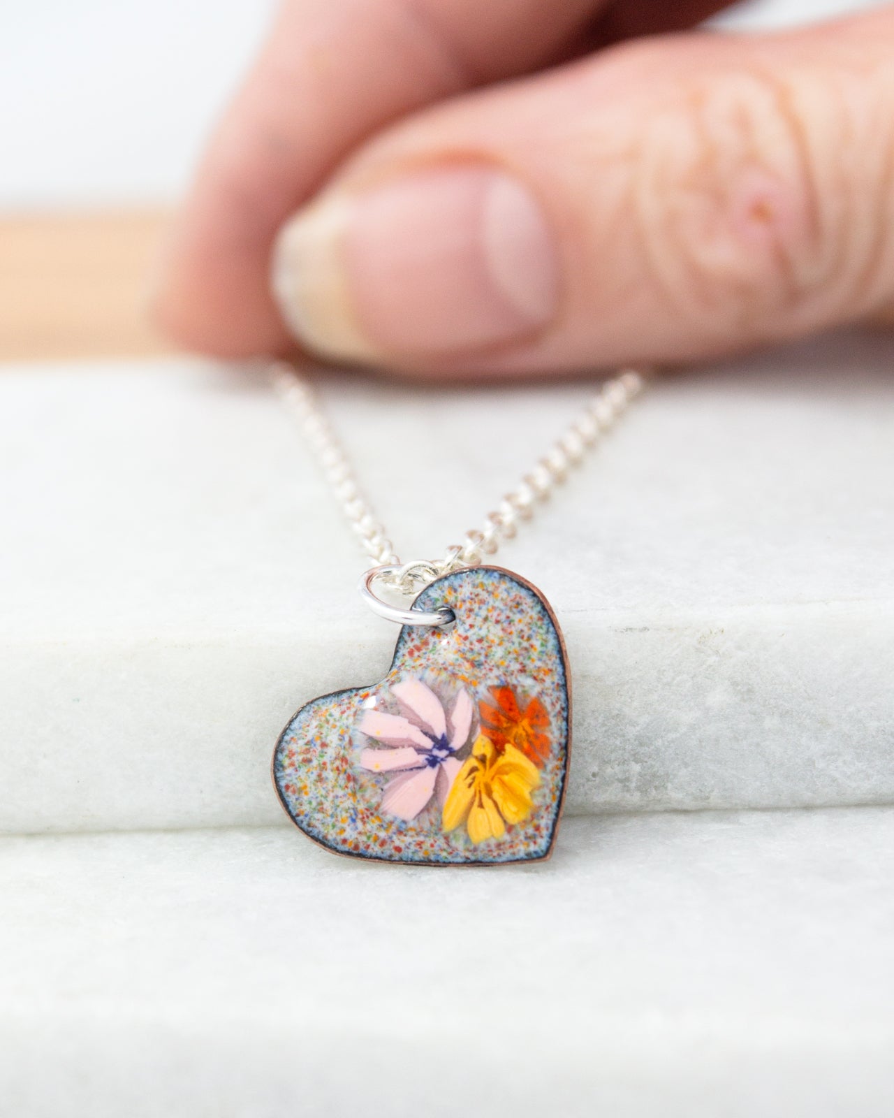 A hand is holding a heart-shaped pendant necklace with a colorful floral design.