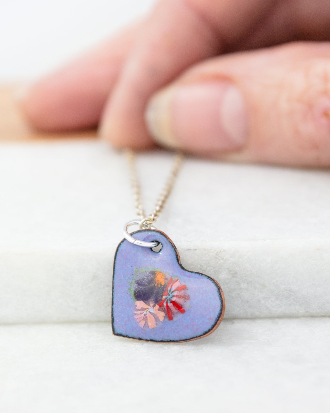 A hand is holding a heart-shaped pendant with a floral design, which is resting on a white surface.