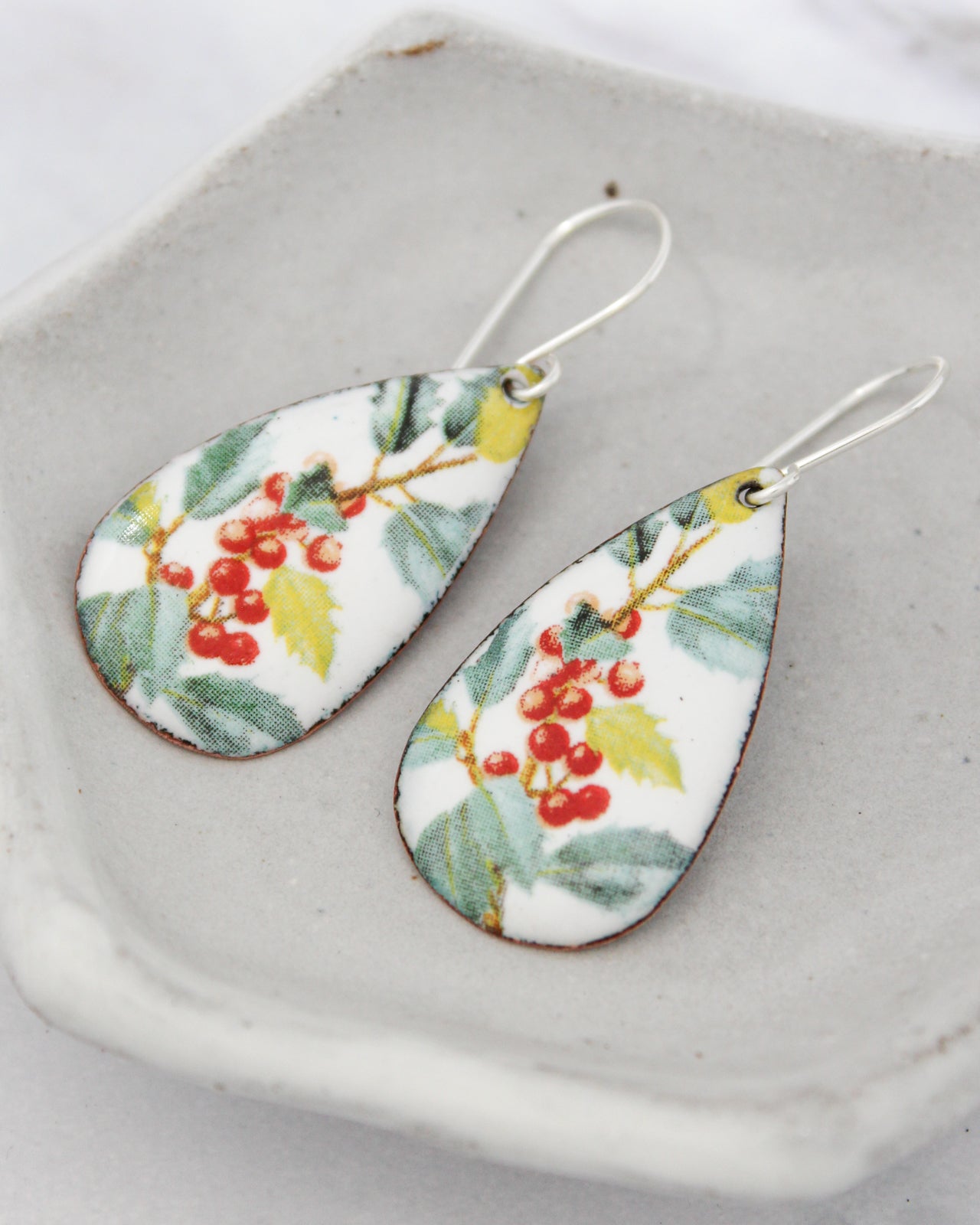 A pair of earrings with a floral design, featuring red berries and green leaves, are displayed on a white plate.