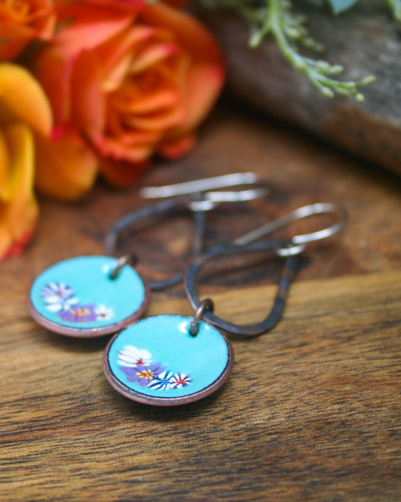 A pair of blue circular earrings with floral designs, placed on a wooden surface next to a bouquet of orange and yellow roses.