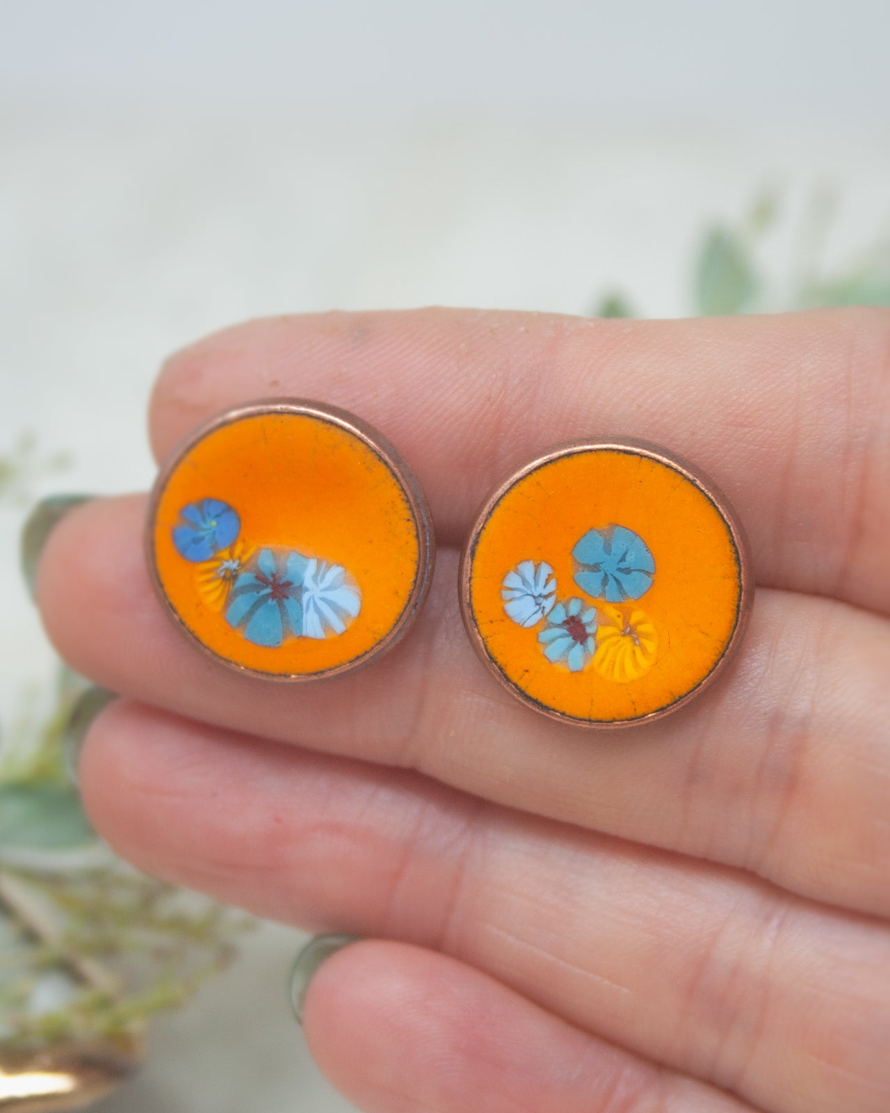 A hand holding a pair of orange earrings with blue and yellow floral designs.