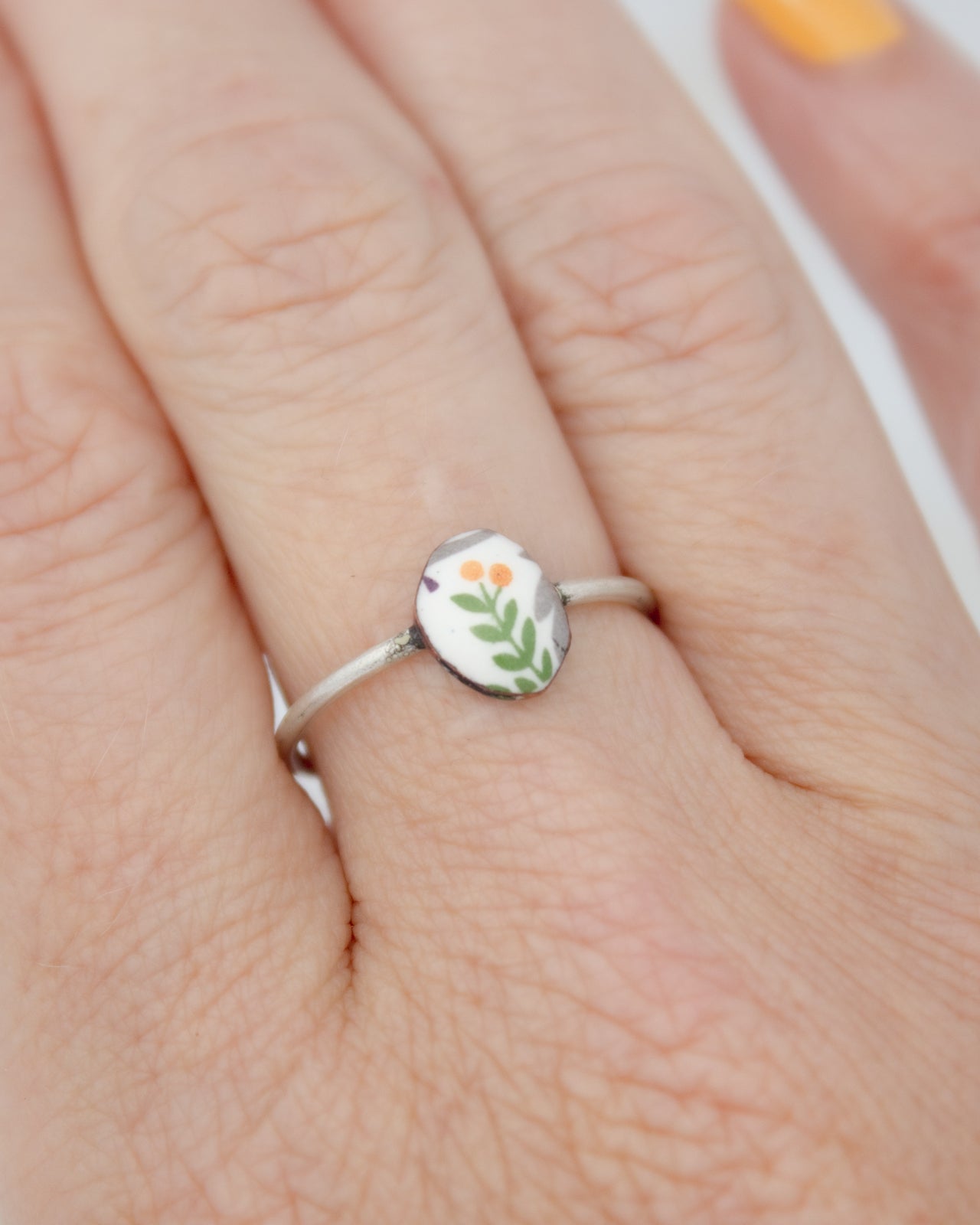 A silver ring with a small oval-shaped design featuring a green leaf and orange flowers.