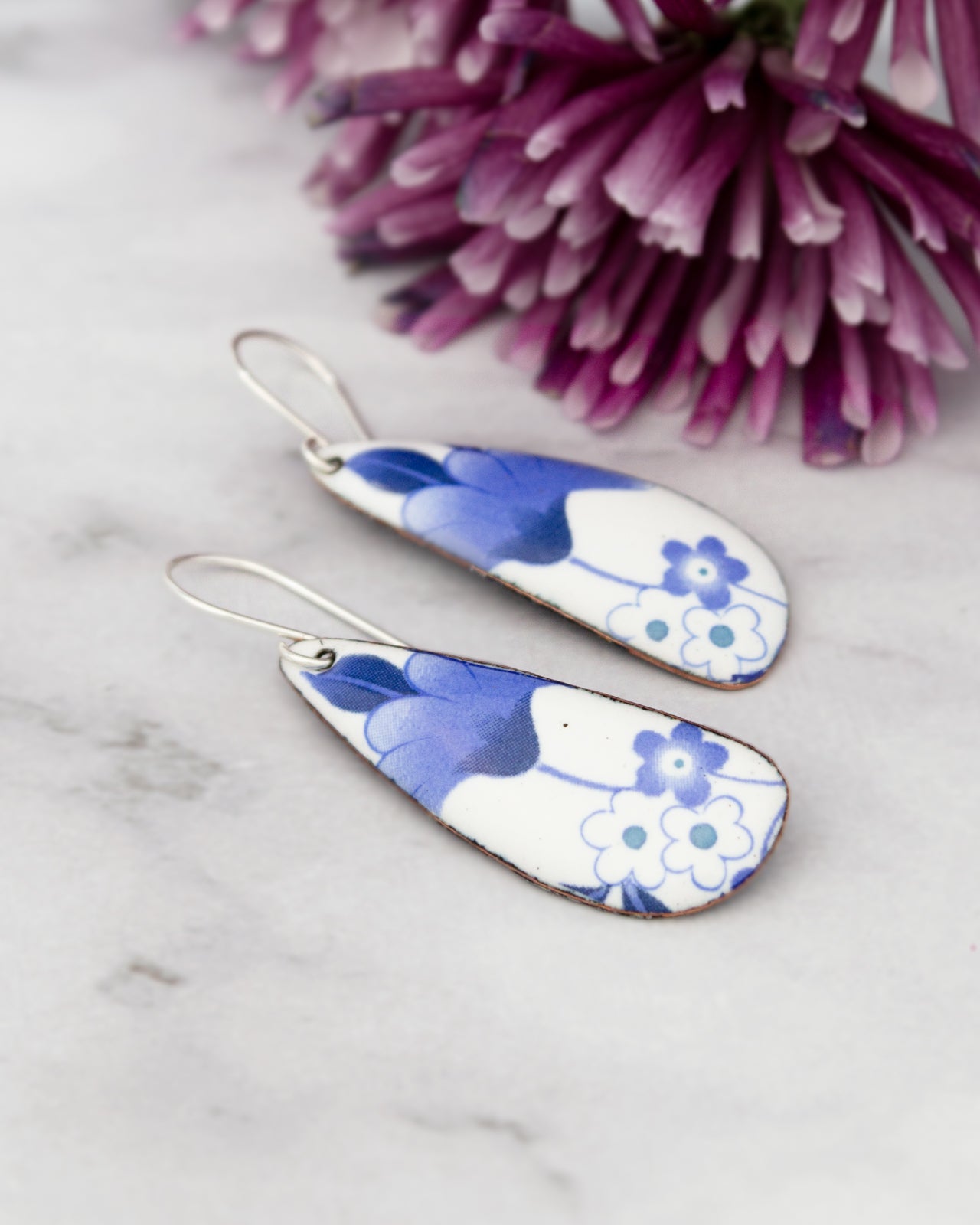 A pair of earrings with blue and white floral designs rests on a marble surface, with a vibrant purple flower in the background.