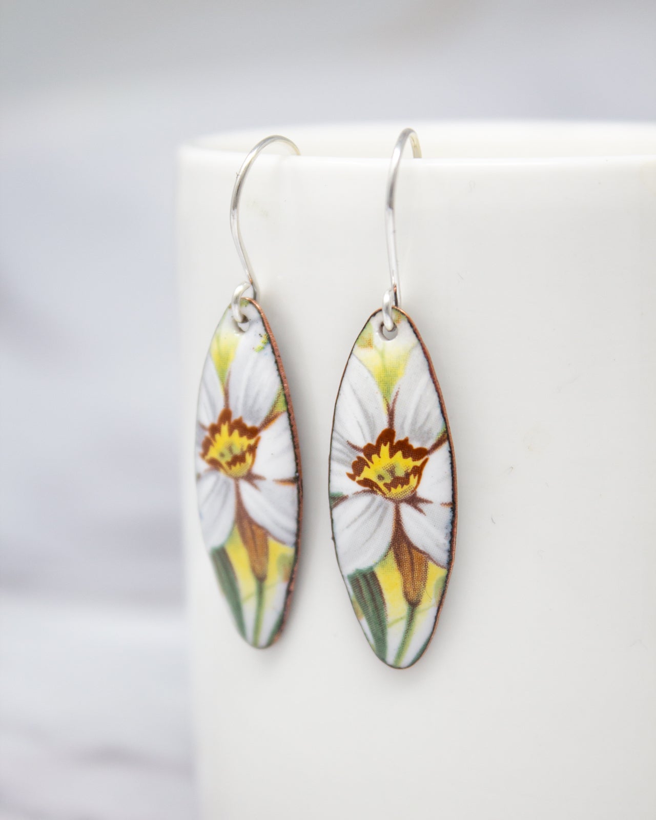 A pair of earrings with a floral design, featuring white and yellow flowers, are displayed on a white surface.